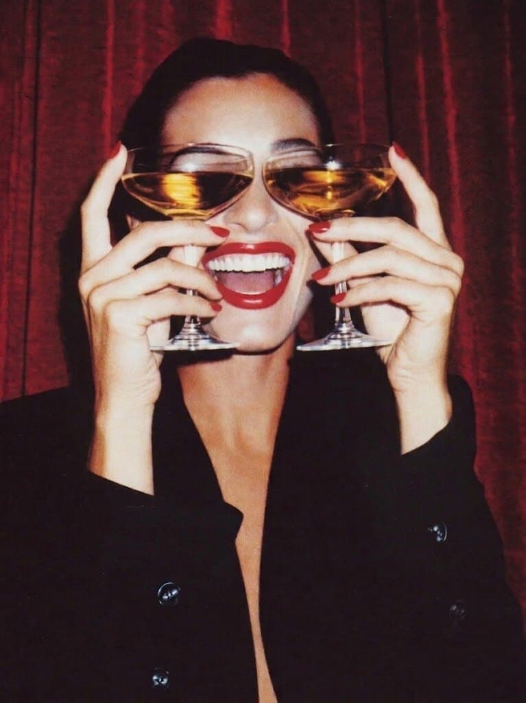 A woman in a black blazer holds two coupes of champagne in front of her eyes against a red curtain